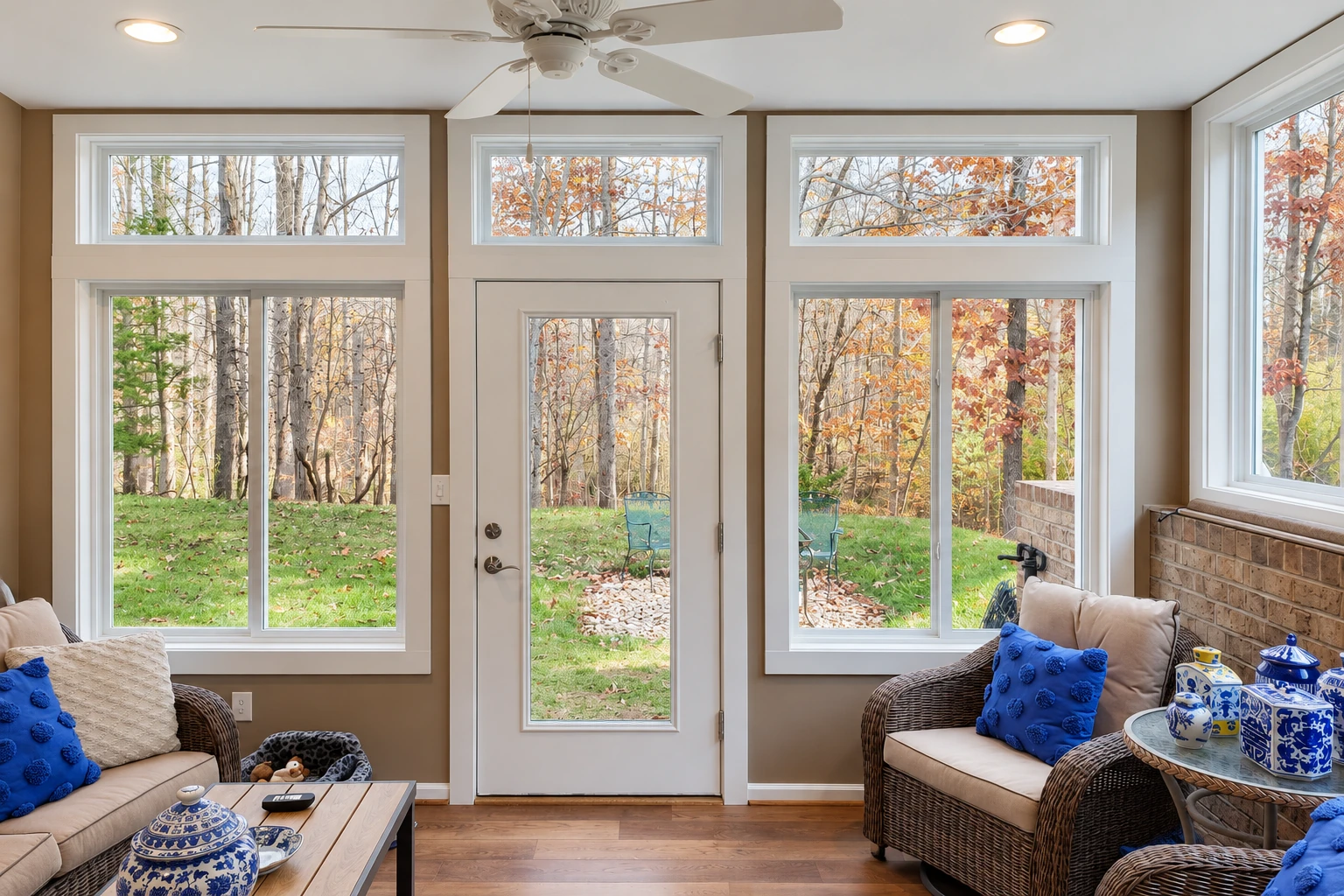 Sunroom converted from a deck, with a central glass door, large windows, and wicker seating facing autumn trees.