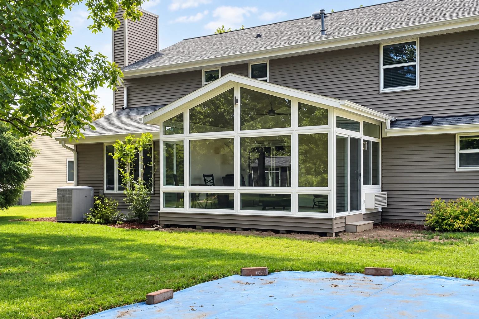 White-framed glass patio enclosure with a peaked roof attached to a gray-sided house.