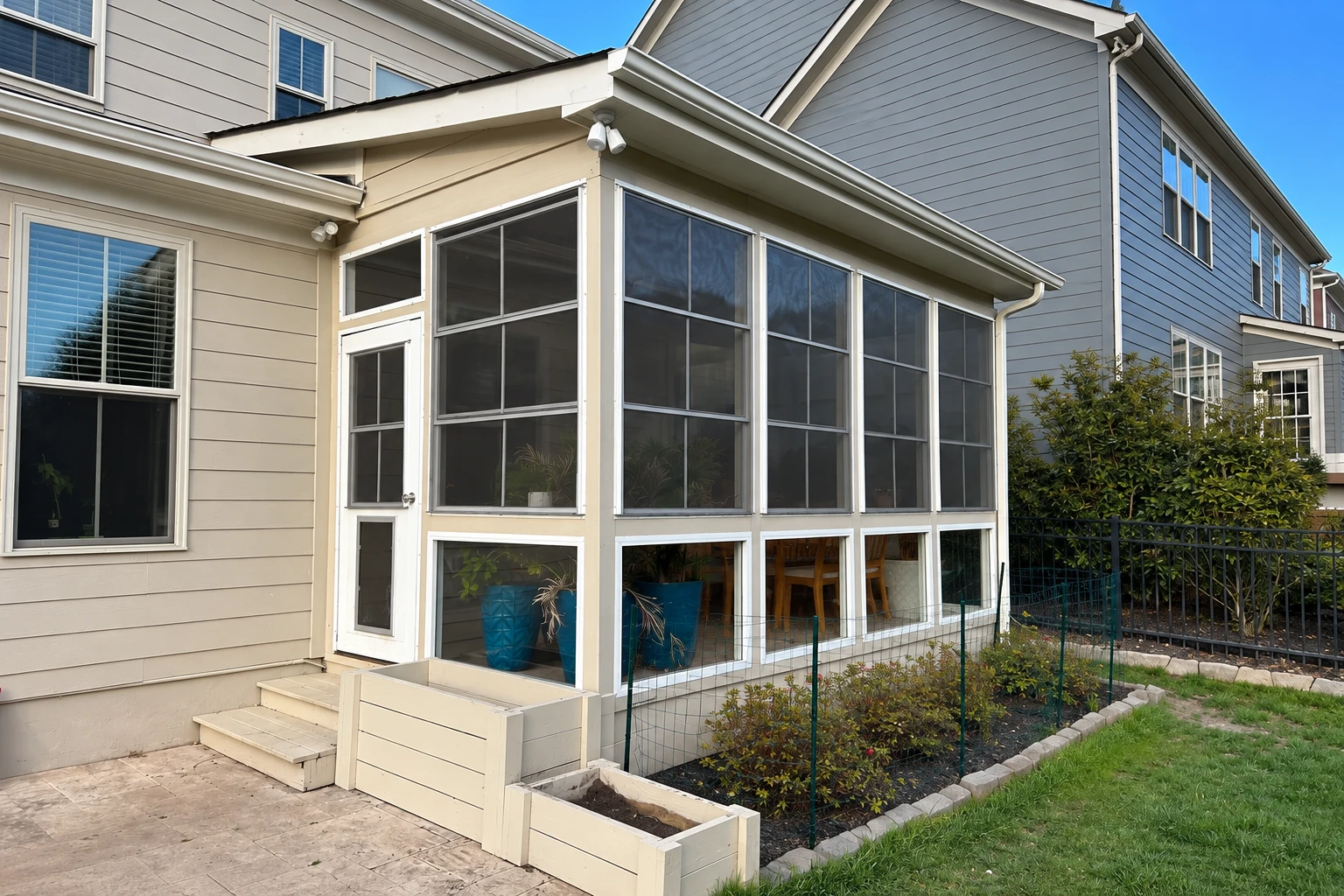 Screened patio enclosure attached to a home, with white framing, a screen door, and garden beds.