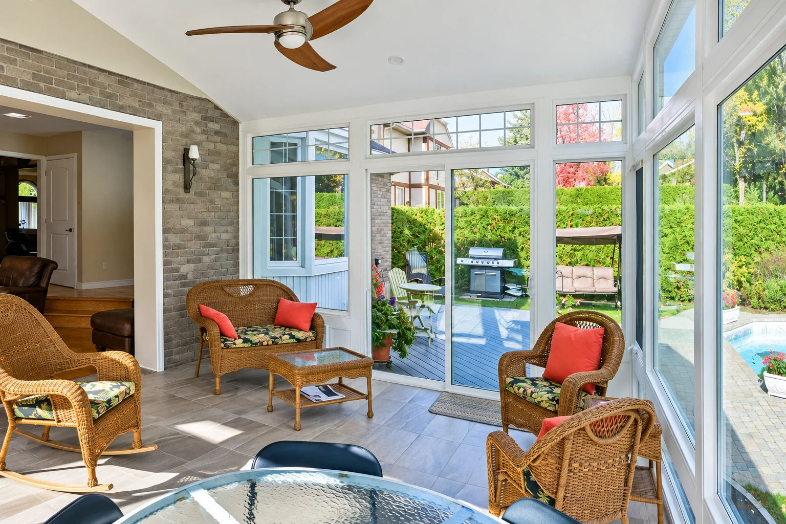 Bright sunroom with wicker seating, tiled floor, and tall windows overlooking a garden and pool.