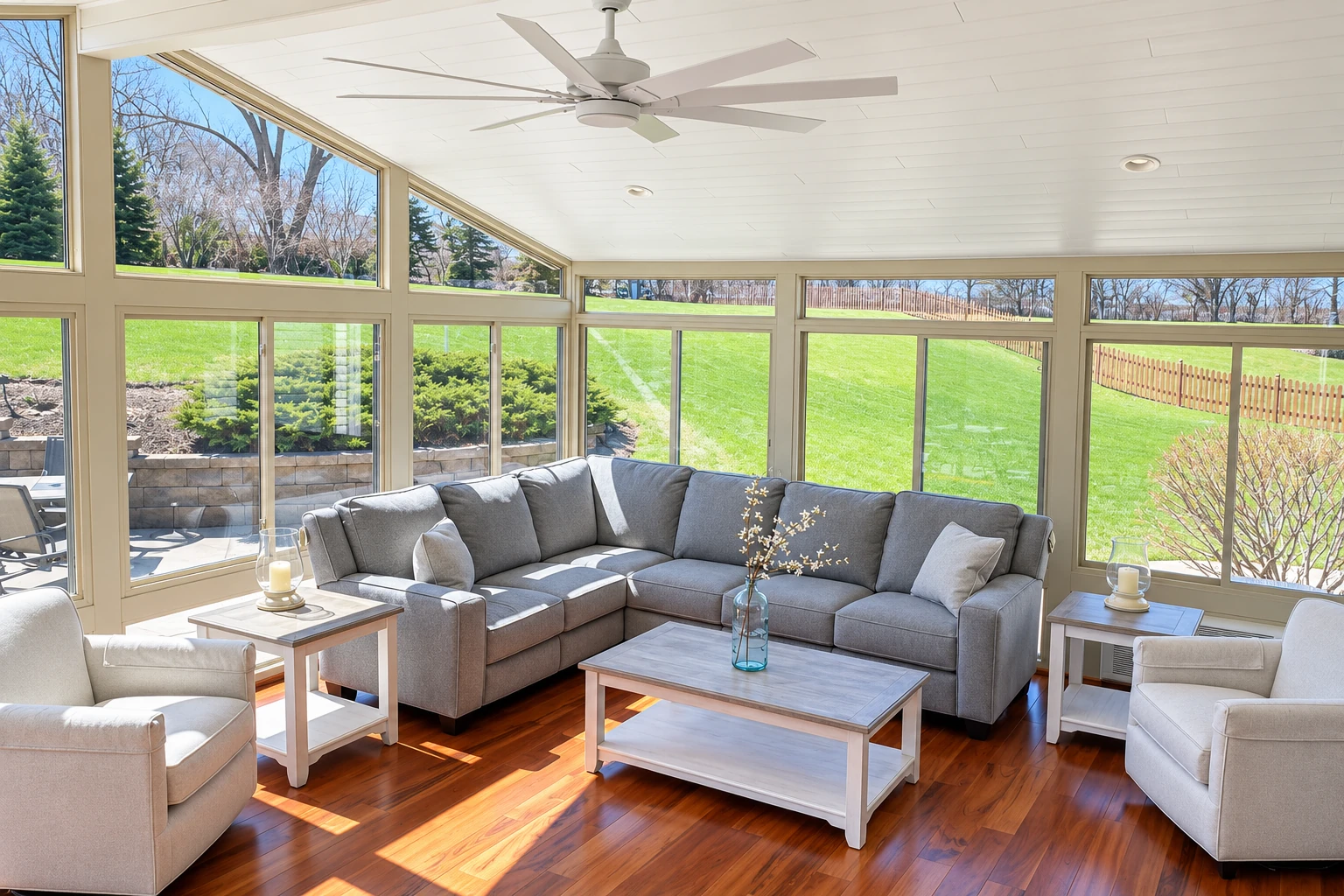 Bright sunroom with a gray sectional, white ceiling fan, and broad windows overlooking a green lawn.